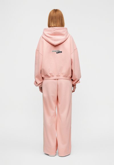 Ροζ φούτερ και παντελόνι φαρδιάς γραμμής από μαλακό ύφασμα. Στο πίσω μέρος έχει μαύρη και τυρκουάζ ετικέτα με γραφικό σχέδιο. Απλή, άνετη εφαρμογή.