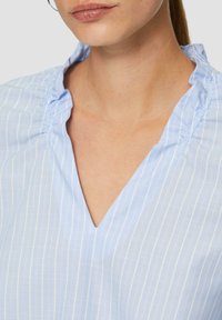 Blouse bleu clair avec des rayures verticales blanches, dotée d'un col en V et de détails froncés près du col. Texture lisse.