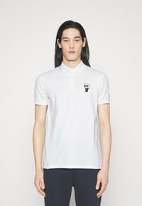 Hvit polo skjorte med korte ermer, to-knappers placket og en svart tegneseriefigur-logo på øvre venstre bryst. Glatt, bomullsmateriale.