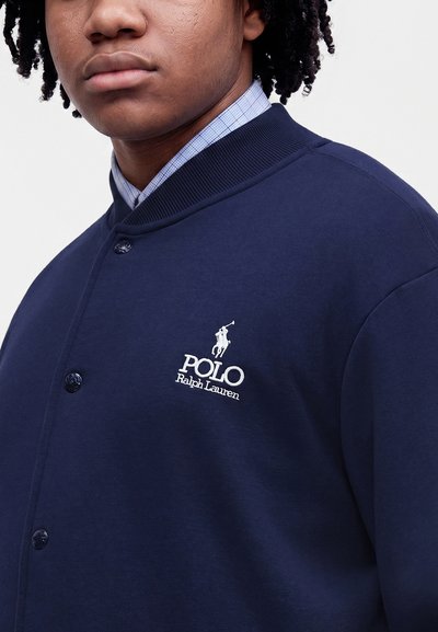Άτομο που φοράει σκούρο μπλε μπουφάν Polo Ralph Lauren με κεντημένο λογότυπο, ορατός γιακάς μπλε καρό πουκάμισου, κοντινή λήψη στο πάνω μέρος του κορμού.