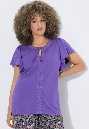 Vrouw met krullend haar die een paarse korte mouwen top met franjes en een broek met patroon draagt, staand met één hand op haar heup.