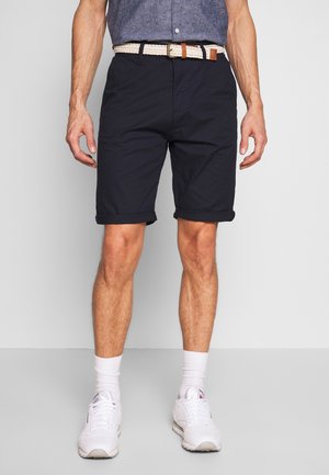 Homme portant un short bleu marine remonté, une ceinture blanche tissée, des chaussettes blanches et des baskets blanches, debout devant un fond uni.