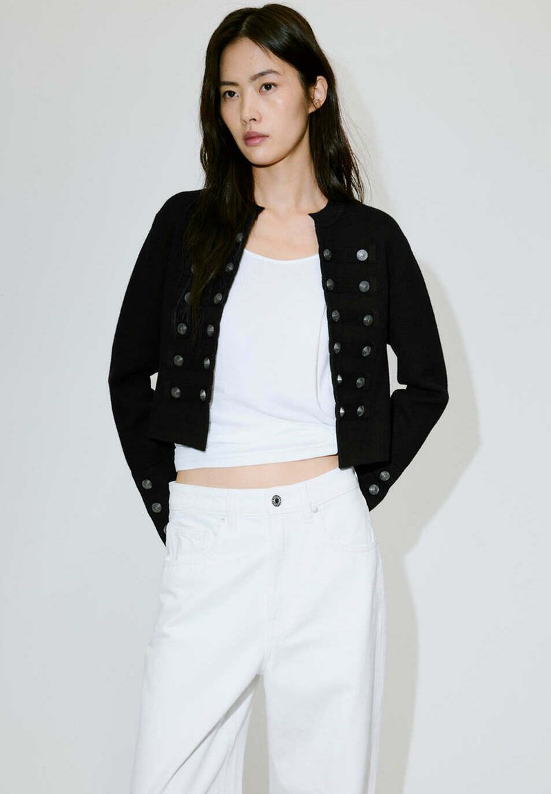 Femme aux longs cheveux foncés porte une veste courte noire avec des boutons sur un haut blanc et un pantalon blanc, debout devant un fond clair uni.