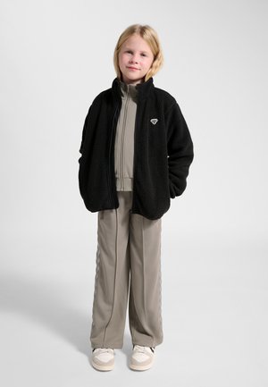 Kind staat met handen in de zakken, draagt een zwarte fleecejas over een grijs zip-up trainingspak en witte sneakers tegen een effen achtergrond.