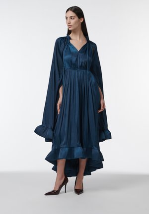 Mujer con vestido midi de satén azul oscuro con bajo con volantes y capa a juego, combinados con zapatos de tacón altos marrón oscuro, de pie contra un fondo blanco.