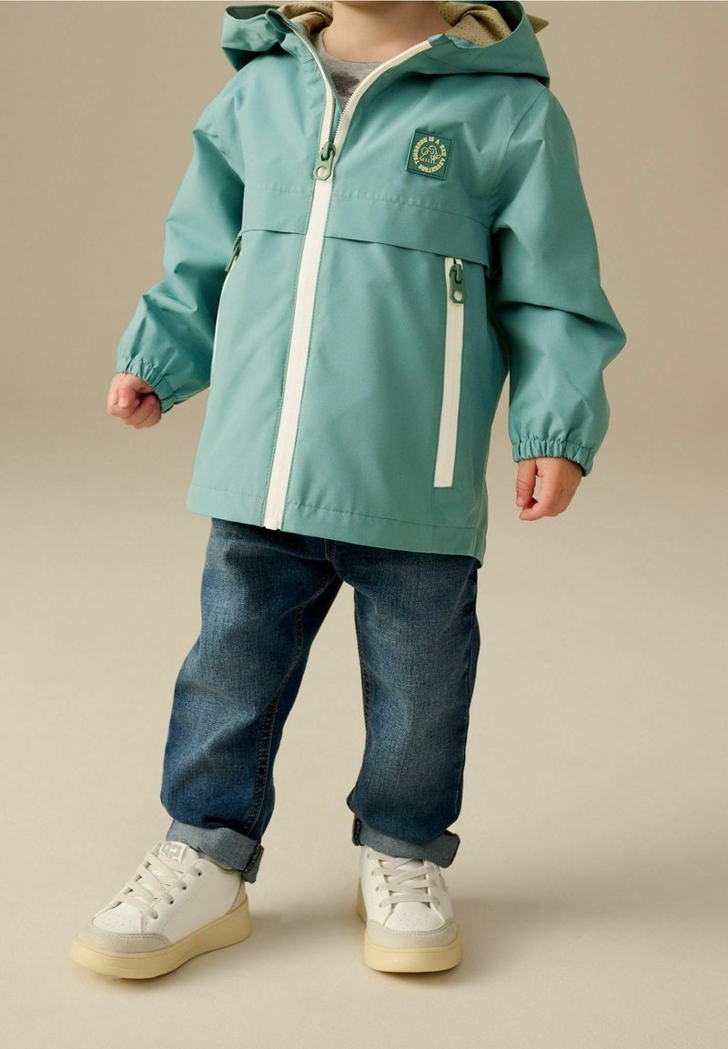 Enfant portant une veste de pluie à capuche turquoise, un jean bleu retroussé et des baskets blanches, debout sur un fond beige.
