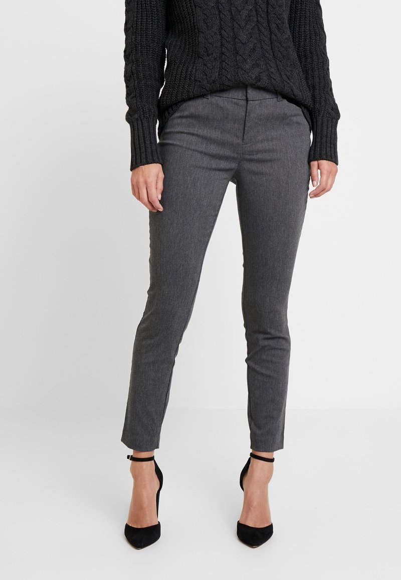 Femme portant un pantalon slim gris, un pull en maille torsadée noir et des talons noirs à bride cheville, debout devant un fond uni.