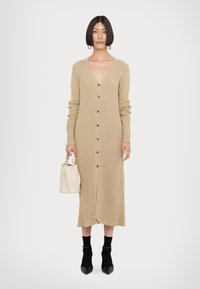 Beige geribbelde gebreide cardigan jurk met lange mouwen, V-hals, knopen aan de voorkant en een rauwe zoom. Gecombineerd met een witte handtas en zwarte enkelsokken.