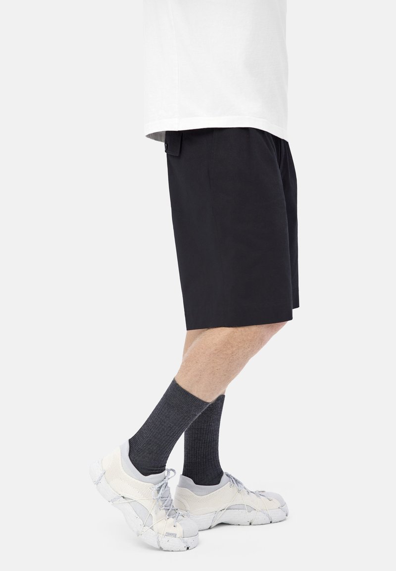 Persona che indossa pantaloni corti neri, calze a coste grigio scuro e sneakers in mesh bianche con suole grigio chiaro, in piedi su sfondo bianco.