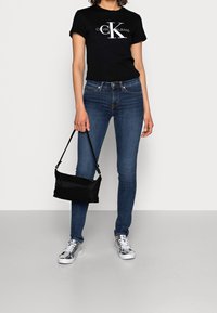 Camiseta de algodón negra con logo blanco, jeans skinny de denim azul, bolso de hombro negro y zapatillas plateadas metálicas. Outfit simple y casual.