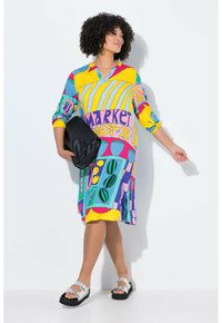 Vestido colorido con una mezcla de patrones en amarillo, azul, rosa y verde, mangas campana y diseño de escote en V; combinado con bolso de mano negro y sandalias plateadas.