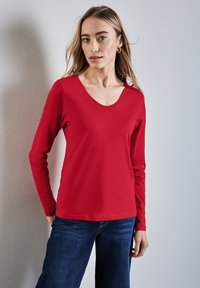 Långärmad, röd bomullsskjorta med v-ringning. Slät textur och figursydd form. Matchad med blå denimjeans.