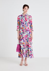 Blommig midi-klänning med knäppning fram, i livliga färger på en ljusgrön bakgrund; matchad med en rosa fransad väska och vita sandaler.