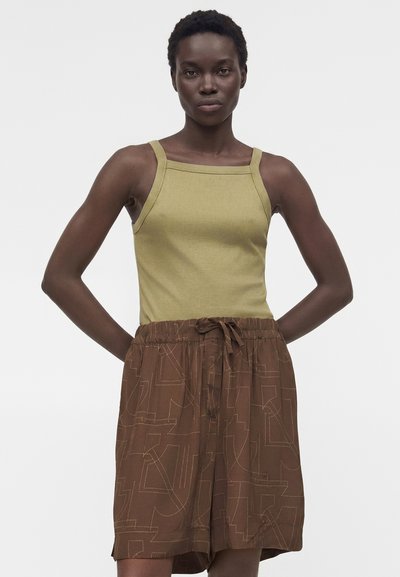 Γυναίκα με κοντά σγουρά μαλλιά που φοράει αμάνικη τοπ ελιάς και χαλαρά καφέ σορτς με γεωμετρικό σχέδιο, χέρια στα ισχία.
