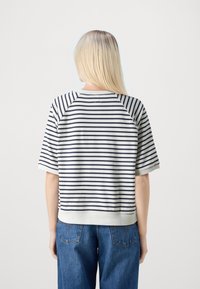 Kvinna med långt blont hår iförd en vit och svart randig kortärmad topp och blå jeans, vänd bort från en enkel vit bakgrund.