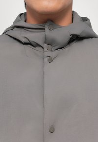 Veste légère grise avec un col haut, une capuche pliée et des boutons-pression sur le devant. Texture lisse avec un design minimaliste.