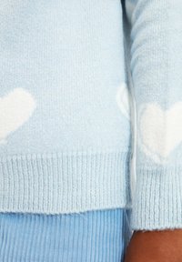 Jasnoniebieski sweter wykonany z miękkiego materiału, z białymi wzorami serc. Dzianina jest prążkowana na mankietach i dolnym brzegu, co dodaje tekstury.