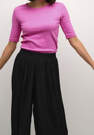 Nainen pukeutuneena istuvaan vaaleanpunaiseen lyhythihaiseen toppiin ja löysiin mustiin korkeavyötäröisiin laskostettuihin housuihin, seisomassa käsivarret hieman ojennettuina.