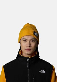 Yellow ribbed beanie with a black logo patch, worn with a black and yellow fleece jacket featuring a high collar and zipper detail.