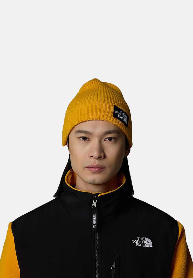 Yellow ribbed beanie with a black logo patch, worn with a black and yellow fleece jacket featuring a high collar and zipper detail.