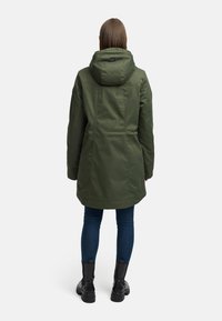 Eine Frau trägt eine olivgrüne Kapuzenparka, dunkle blaue Skinny-Jeans und schwarze Stiefeletten und steht mit dem Rücken zu einem weißen Hintergrund.