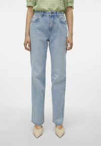 Ανοιχτό μπλε ψηλόμεσα τζιν με ίσια πόδια, ορατές ραφές στις μπροστινές τσέπες και ελαφρώς ξεθωριασμένο υφή, σε συνδυασμό με κρεμ καμπυλωτά παπούτσια.