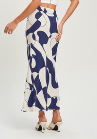 Gonna lunga e aderente color crema con motivi astratti blu navy, dotata di una fascia in vita elasticizzata e una fessura laterale. Indossata con sandali con i piedi scoperti.