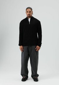 Zwarte geribbelde sweater met ritssluiting en hoge kraag, gecombineerd met loszittende donkergrijze denim broek en zwarte schoenen met roze accenten.