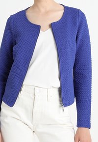 Blå strukturerad zip-up jacka med rund halsringning, ihopparad med en vit blus och ljusa byxor. Tyget har ett mönstrat mönster.