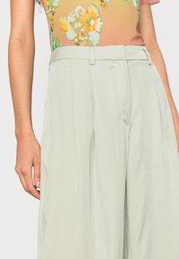 Calças largas verde-claro com uma textura suave, apresentando pregas e uma cintura alta, complementadas por um brilho subtil.