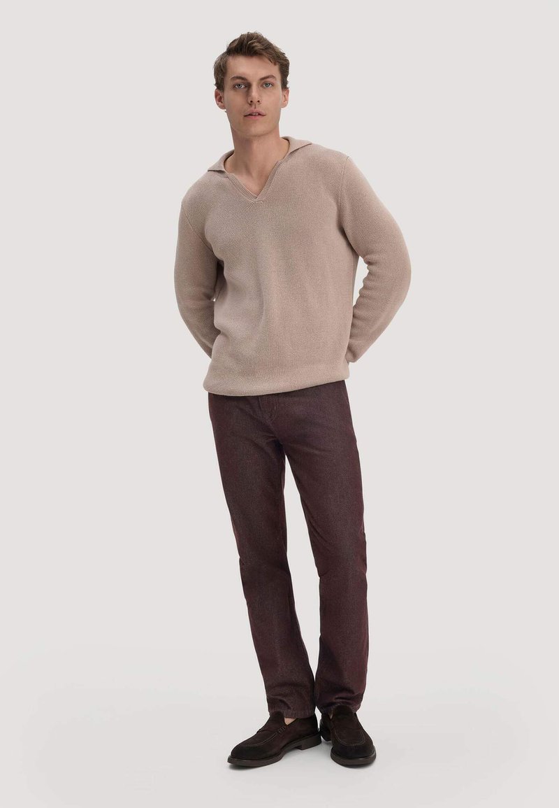 Jeune homme debout, portant un pull en maille beige, un pantalon marron foncé et des chaussures noires sur un fond clair uni.