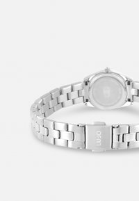 Montre-bracelet en métal argenté avec un design à maillons, un boîtier rond et un dos transparent. Fermée avec un fermoir portant le nom de la marque.