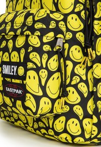 Černý batoh s žlutým vzorem usměvavých obličejů. Obsahuje přední kapsu, logo Eastpak a černé šňůrky se žlutými konci. Texturovaná tkanina.