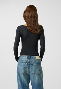 Schwarzer langärmeliger Body, kombiniert mit blauen hoch taillierten Jeans aus Denim, die zwei Gesäßtaschen und ein sichtbares Lederpatch am Bund aufweisen.