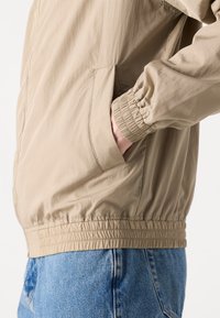 Béžová lehká bunda s hladkou texturou, elastickými manžetami a pasem. Má boční kapsy a volný střih.