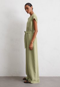 Tuta verde oliva, con gambe ampie in tessuto leggero, maniche corte, una tasca frontale e una cintura in vita per una vestibilità regolabile.