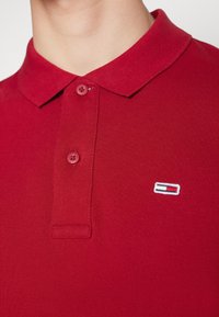 Červená polo košile s texturovaným pletem, s klasickým límečkem, dvěma knoflíky a malým vyšitým logem v červené, bílé a modré barvě.