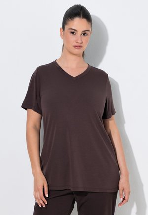 Donna con capelli scuri legati indossa una maglietta semplice marrone scuro a maniche corte con scollatura a V e pantaloni coordinati, in piedi contro un muro bianco.