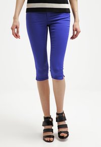 Pantalones capri azules elásticos con corte slim, que presentan un dobladillo deshecho. Combinados con sandalias negras de tacón alto con tiras sobre un fondo claro.