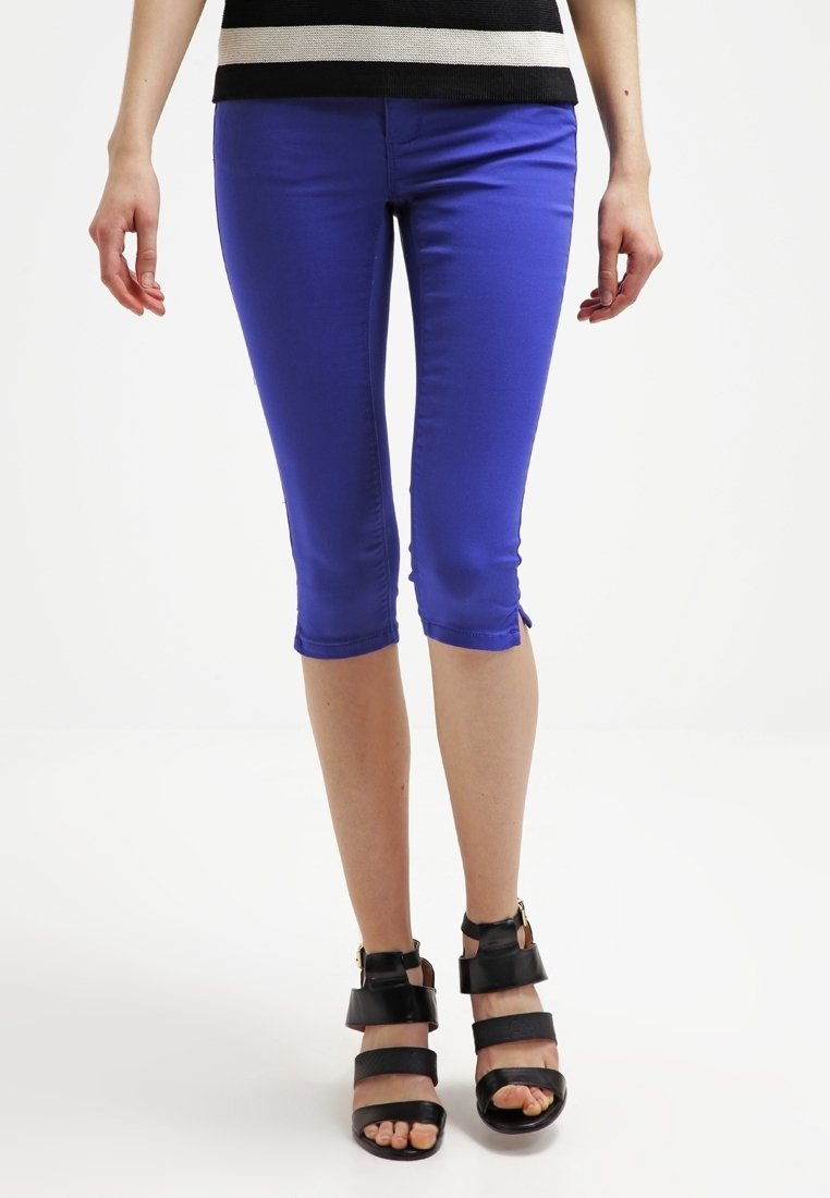 Pantalones capri azules elásticos con corte slim, que presentan un dobladillo deshecho. Combinados con sandalias negras de tacón alto con tiras sobre un fondo claro.
