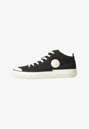 Baskets basses en toile noire avec une semelle en caoutchouc blanche, un bout renforcé, des lacets blancs et un écusson rond blanc avec logo sur le côté.