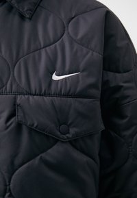 Black quilted jacket with a smooth texture, featuring a pointed collar, snap button, and a white Nike logo on the chest.