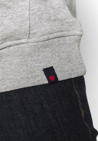 Grijze sweatshirtstof met ribbelstructuur, voorzien van een marineblauw label met een rode cirkelaccent aan de zoom, gecombineerd met donkere denim.