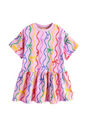 Jong meisje staat met gespreide benen, draagt een roze jurk met kleurrijke lintpatronen, witte sneakers en regenbooggestreepte sokken op een beige achtergrond.
