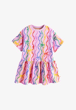 Jong meisje staat met gespreide benen, draagt een roze jurk met kleurrijke lintpatronen, witte sneakers en regenbooggestreepte sokken op een beige achtergrond.
