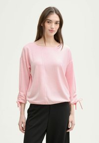 Gestreifter, hellrosa Pullover mit weitem Ausschnitt und Dreiviertelärmeln. Enthält geraffte Akzente mit Bindebändern an den Ärmeln. Kombiniert mit schwarzen Hosen.