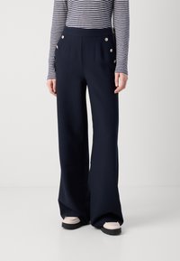 Pantalons larges bleu marine avec accents à double bouton, design plissé et texture lisse, associés à un haut à rayures blanches et noires à manches longues.
