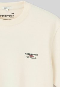 T-shirt en coton de couleur crème avec un col rond, des manches courtes et un graphique sur le devant avec "Paddington aime la marmelade" en texte noir et rouge.