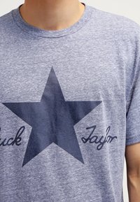 Šedá hezká krátkosrstá tričko s tmavě modrou hvězdou a textem "Chuck Taylor" dole v kurzívě. Měkká tkanina.