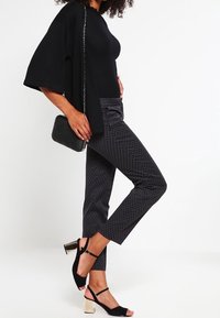 Haut noir côtelé, pantalon foncé à motif, et escarpins noirs avec bride à la cheville et talon bloc blanc. Le mannequin tient un petit sac à main noir.